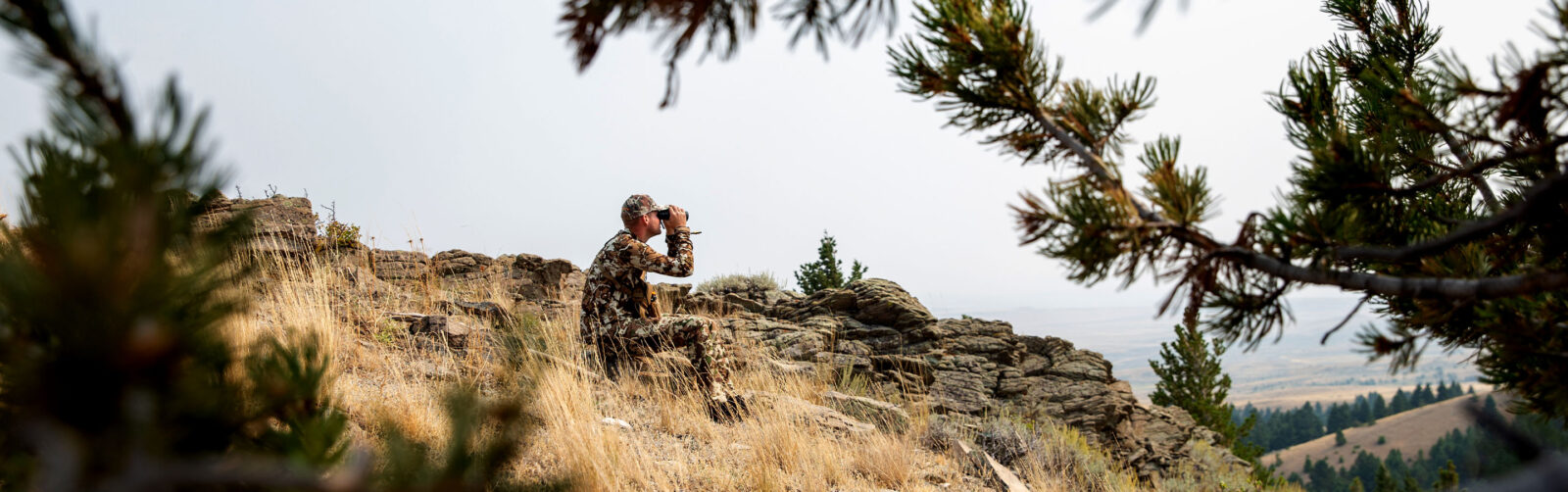 A man sitting on a ridge using binoculars.