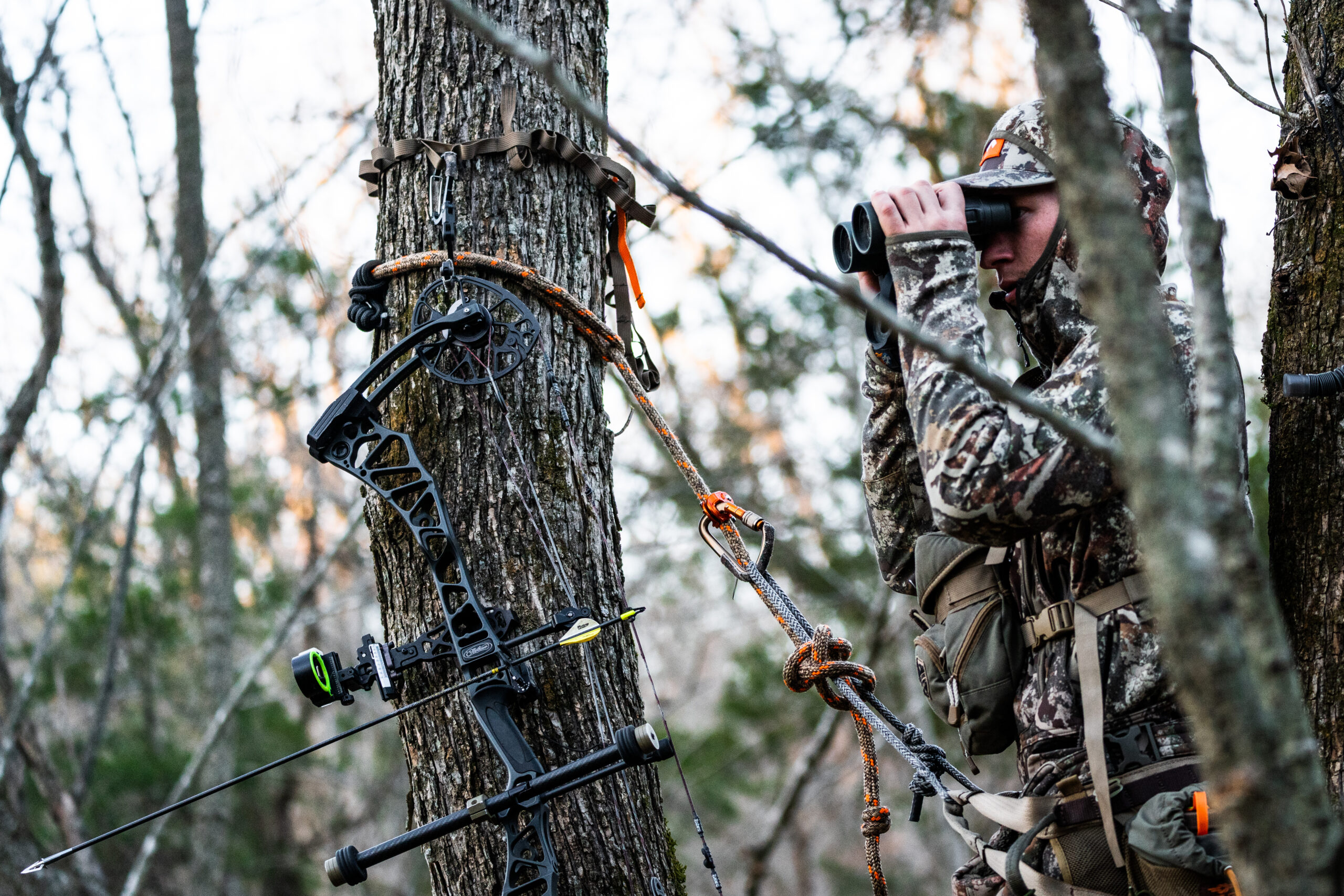 a hunter in full camo in a tree saddle with binoculars and a bow 
