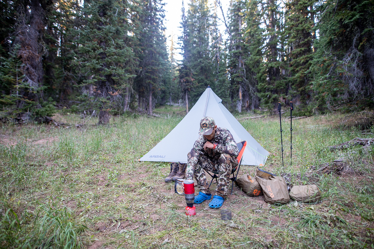 A hunter uses a jet boil sitting in an ultralight chair with an Argali tent behind him. 