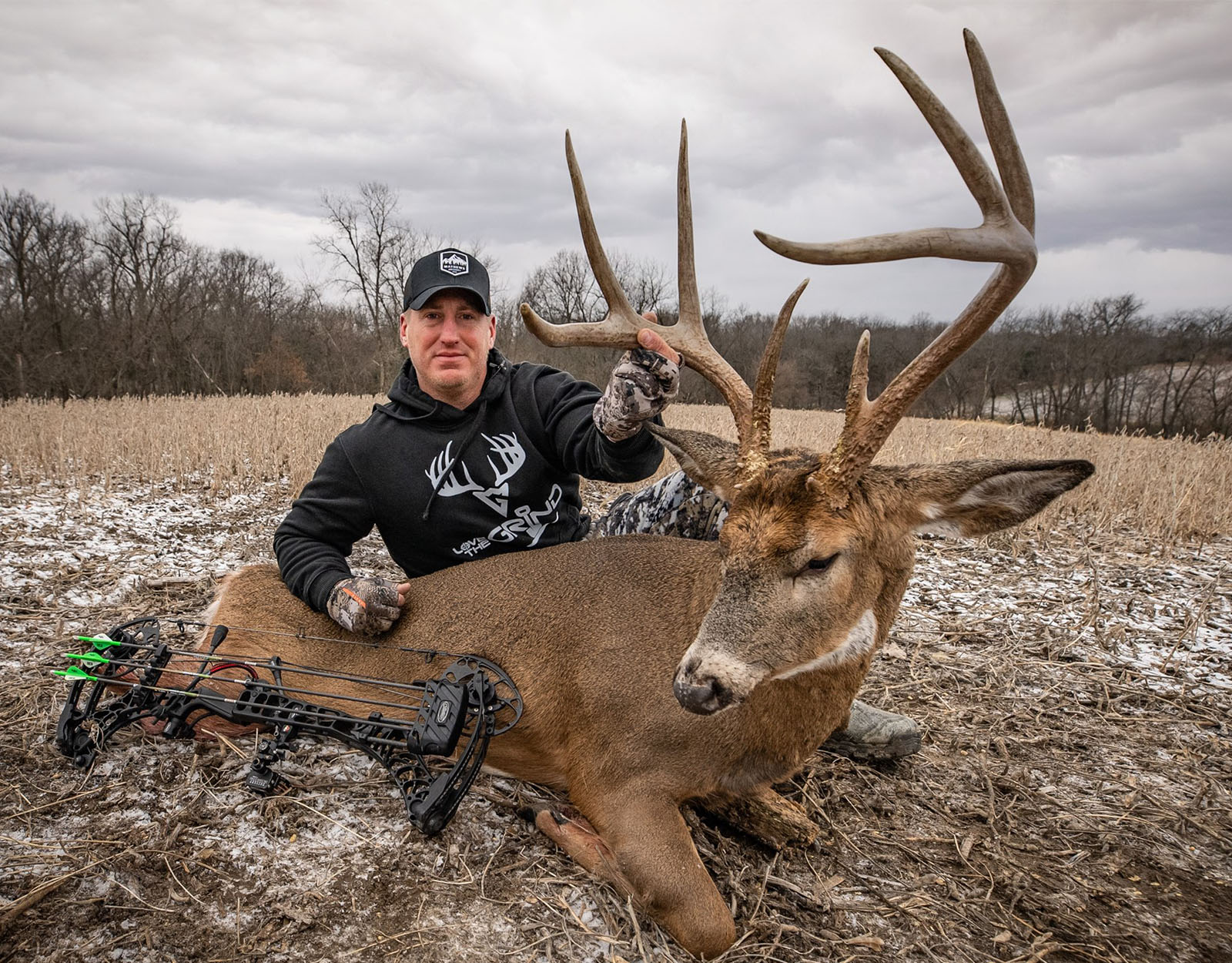 A hunter with a large buck he successfully harvested, with a bow placed across the deer, in a snow-dusted field.