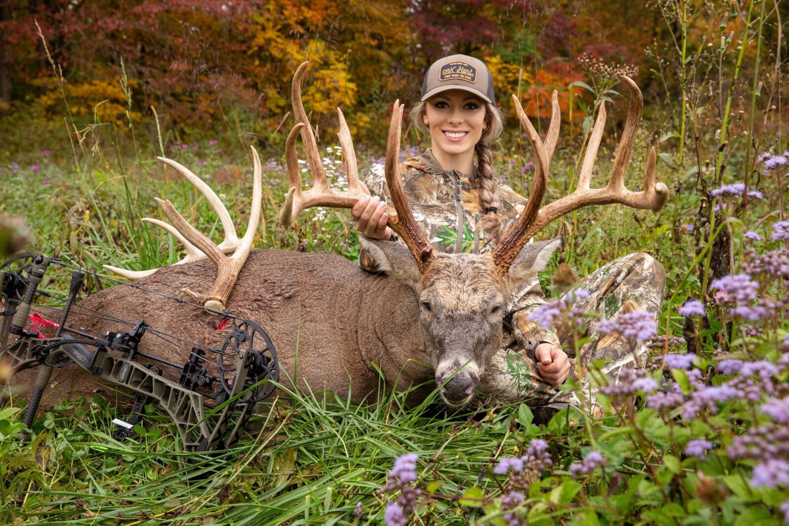 A female hunter with a large buck she successfully harvested, with a crossbow laying in front of the deer, in a field of wildflowers.