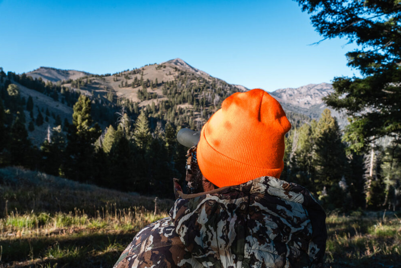 Man in camouflage and blaze orange hat hunting in mountains.