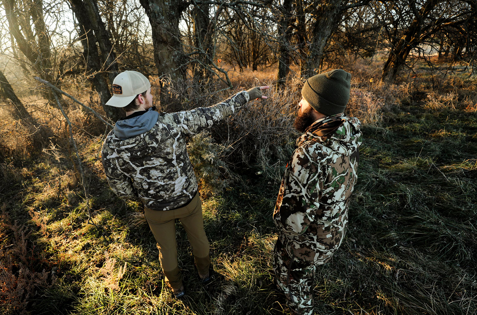 Two men wearing camo stand next to one another in the woods. One man points at something in the distance.