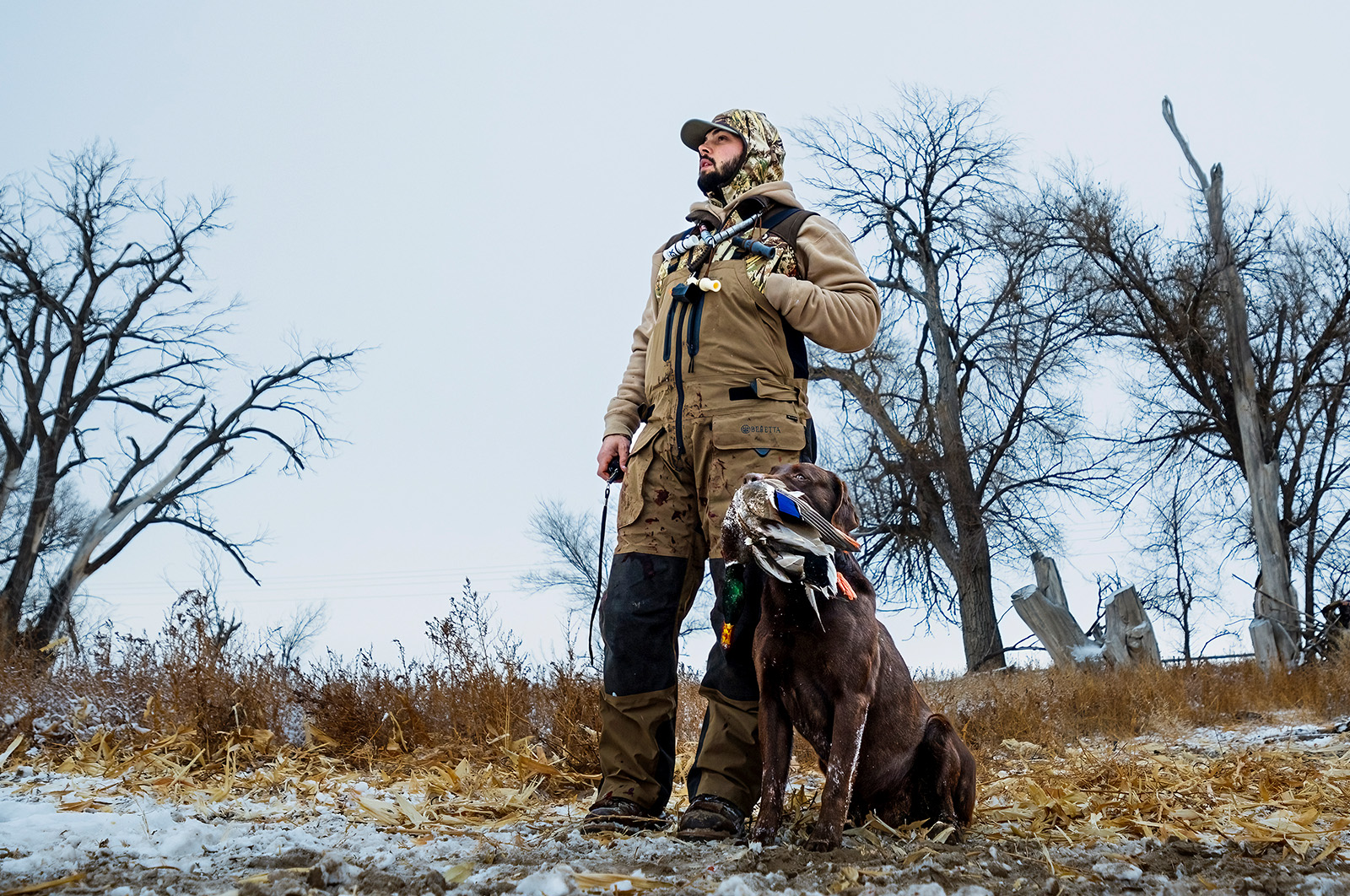 A waterfowl hunter stands with his dog who is holding a duck in its mouth.