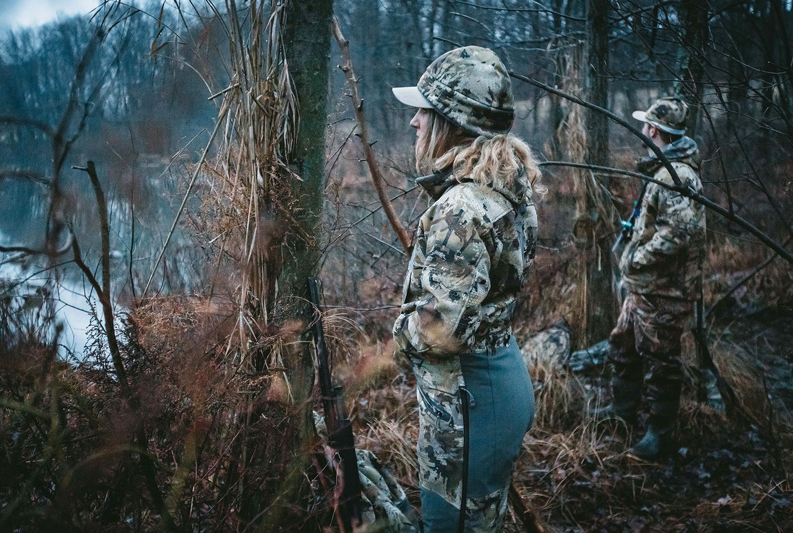 Two hunters look out onto a waterway. 