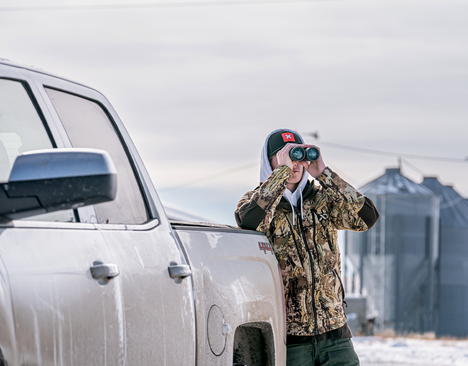 A hunter stands next to a white pickup truck and uses binoculars to scout. 