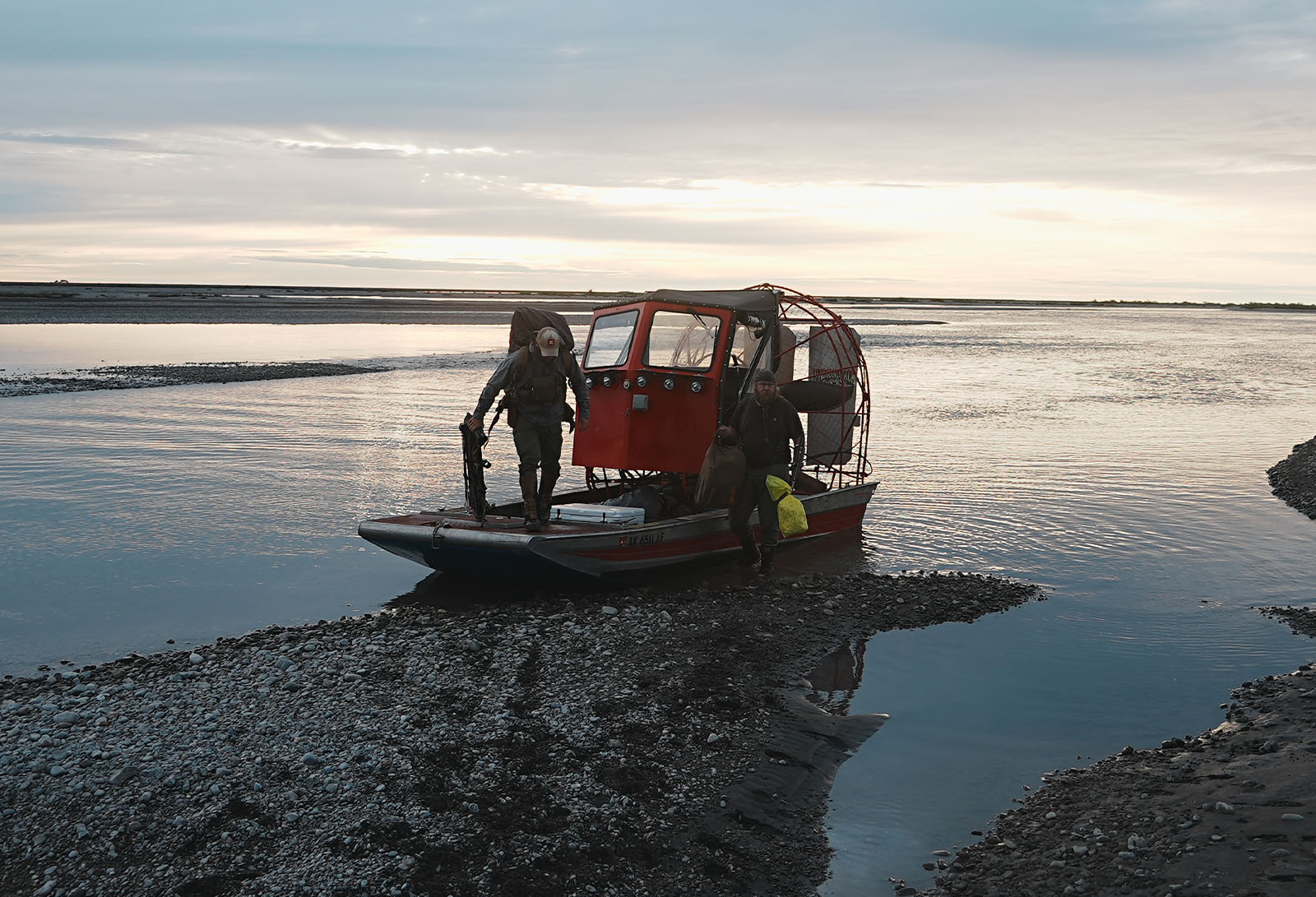 Two hunters disembark from a fan boat onto a rocky shore. 